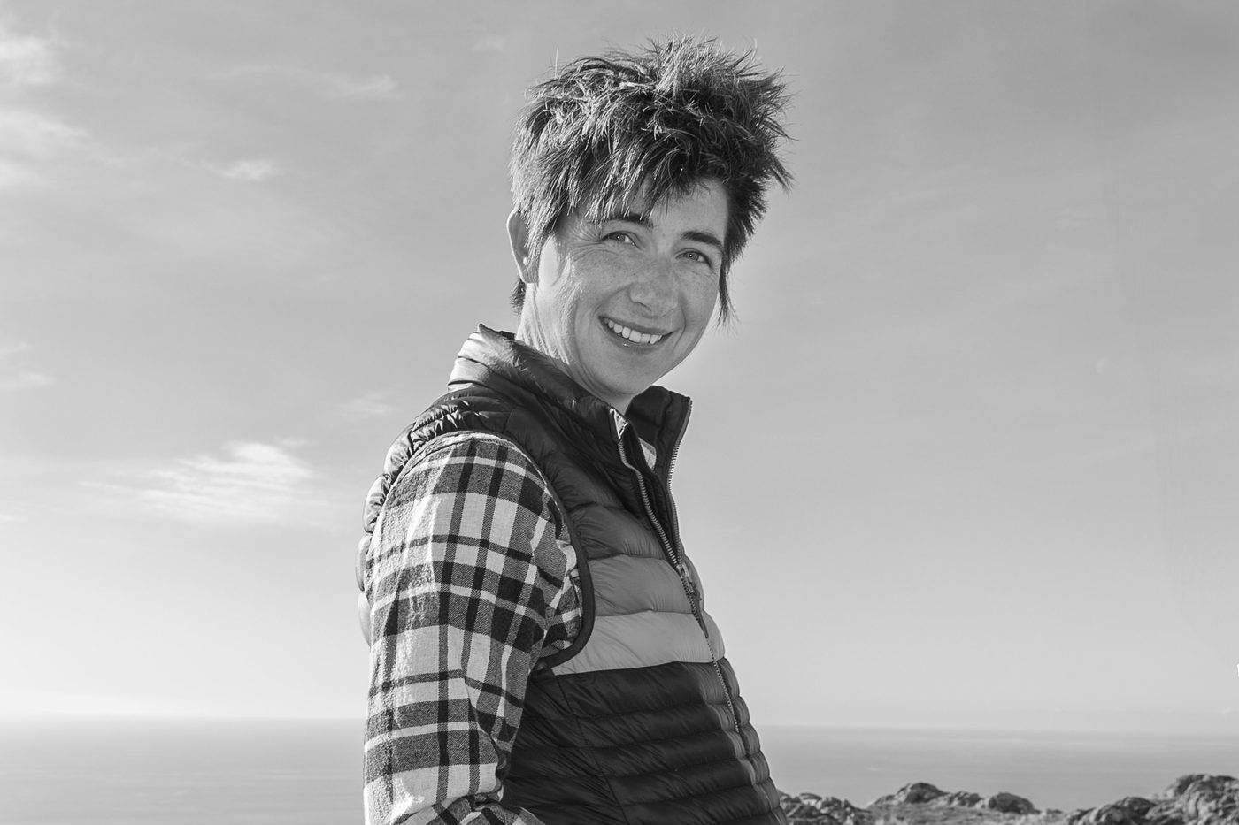 Poppie Johnson smiling over her shoulder, in a checked shirt and puffer vest, on a hilltop above the sea.
