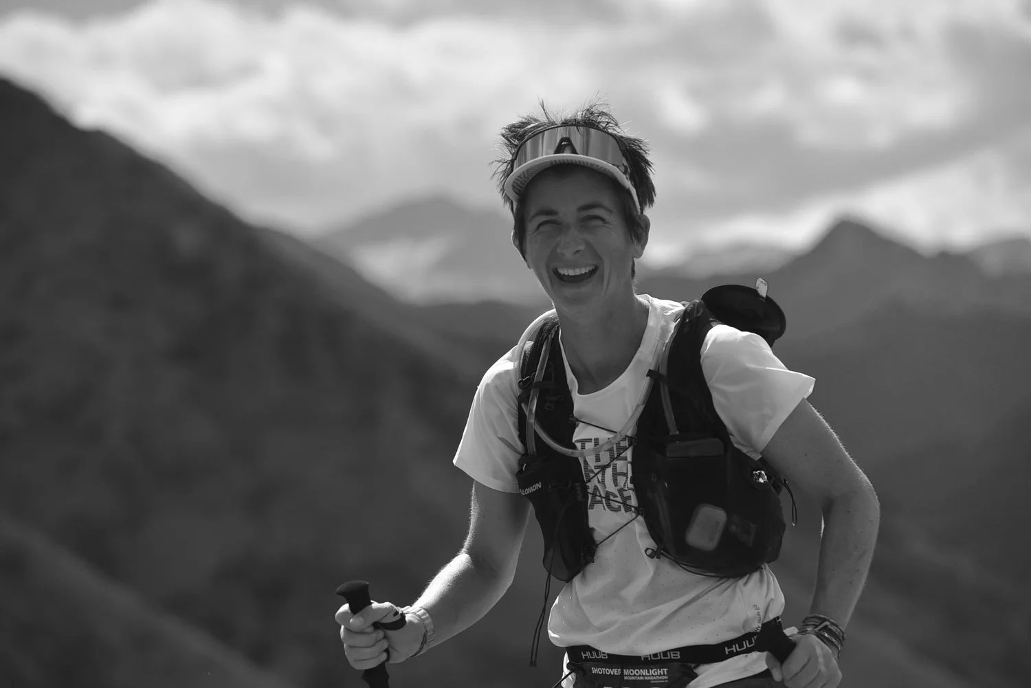 Poppie Johnson smiling, in running gear, sunglasses on her forehead, holding trekking poles, outdoors with mountains and sky in the background.