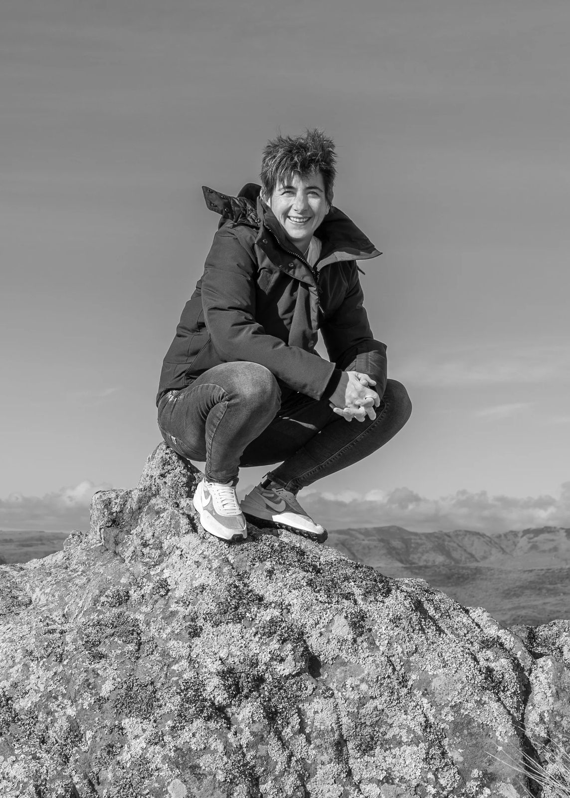Poppie Johnson smiling, crouched on a large rock with hills and a cloudy sky behind her.