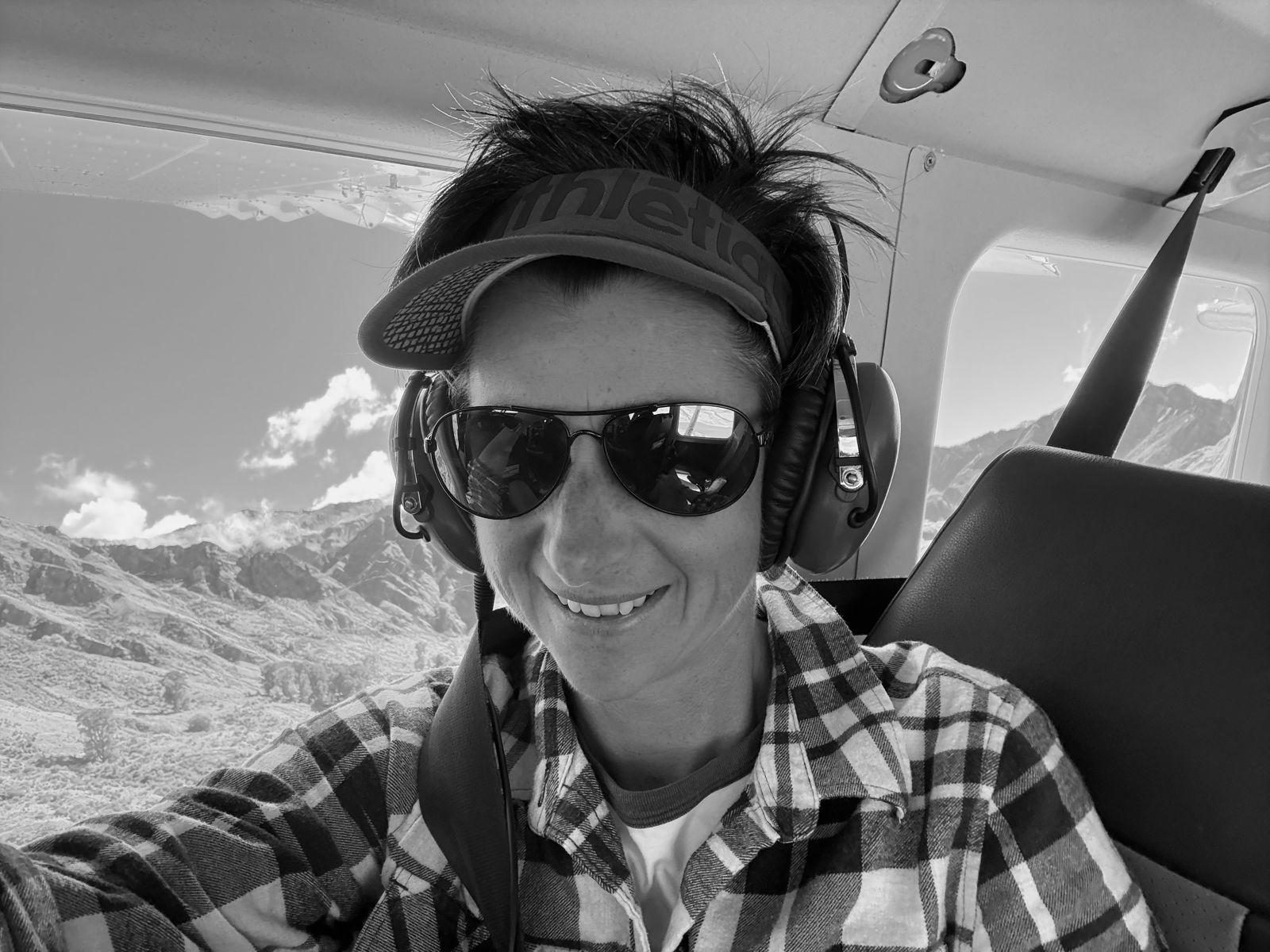 Poppie Johnson in a light plane's front seat wearing a headset and sunglasses, with brown high country hills below