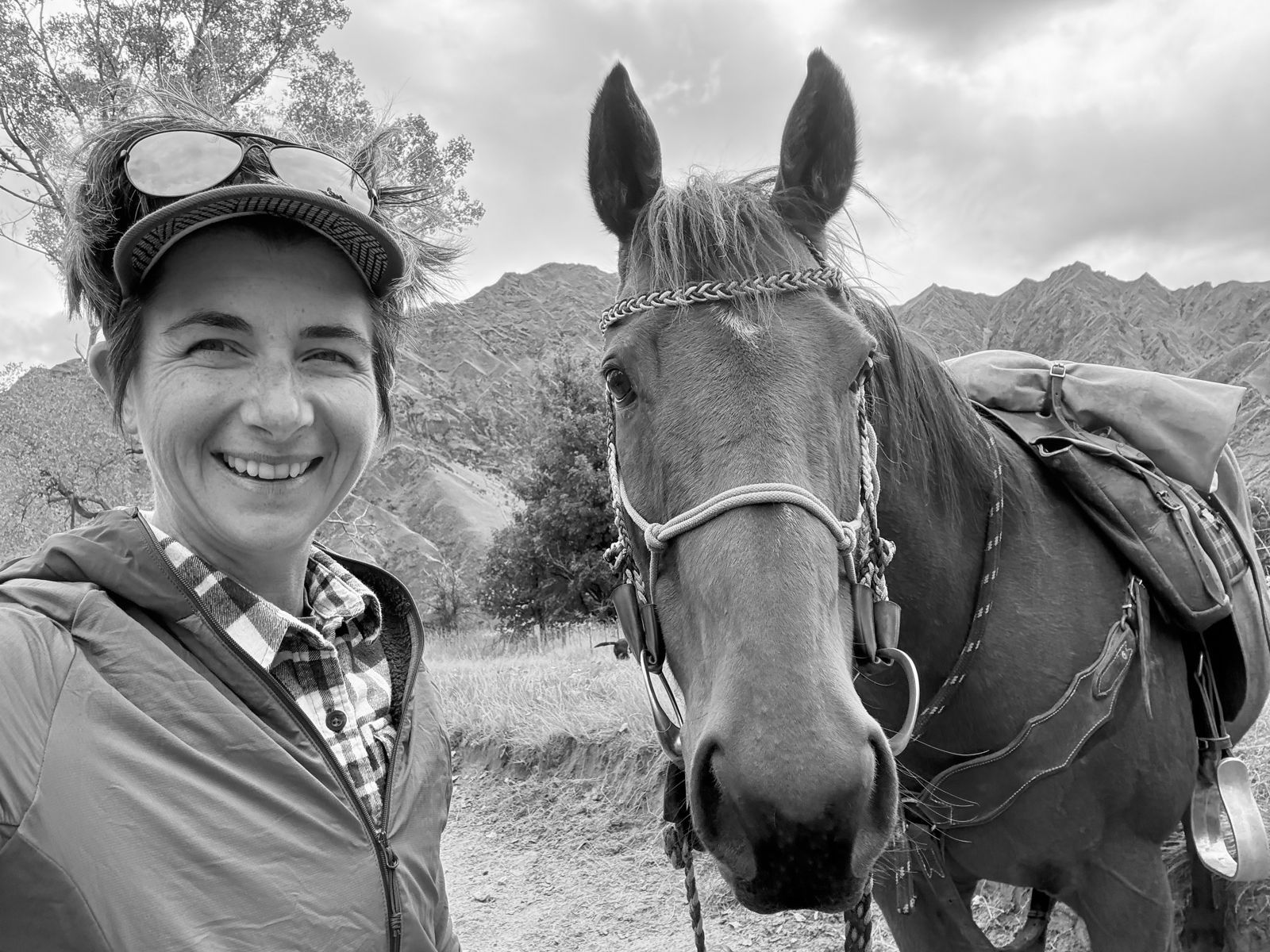 Poppie Johnson smiling beside a saddled packhorse on a dirt track, with mountains behind