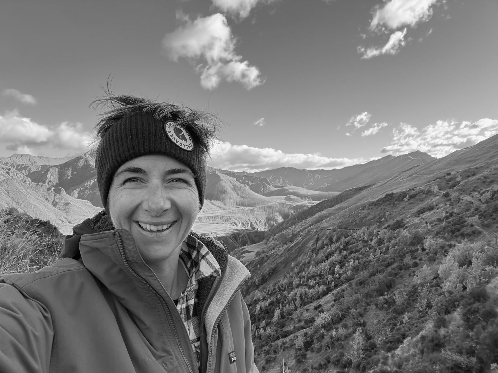 Poppie Johnson smiling in a beanie and blue jacket on a ridge, with high country valleys and mountains behind